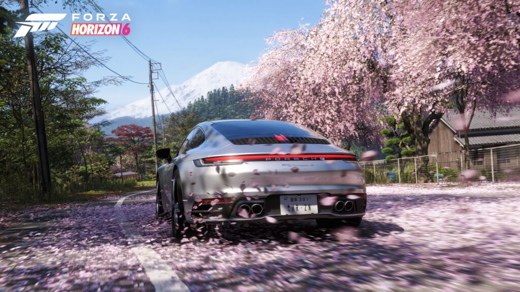 A silver Porsche 911 Carrera S driving down a rural Japanese road covered in fallen pink cherry blossom petals, with a snow-capped Mount Fuji in the background.